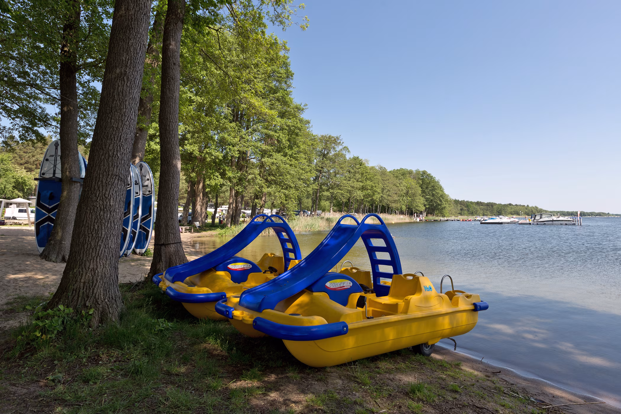 Zwei gelb-blaue Tretboote mit Wasserrutsche am Ufer des Jabelschen Sees, daneben SUP-Boards unter Bäumen